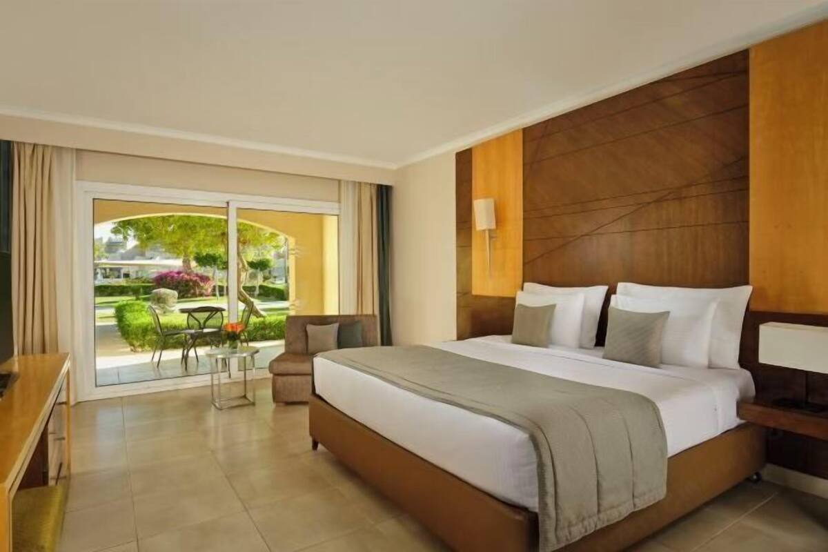 Habitación de hotel con una cama grande, un escritorio y vista a un jardín por una puerta corredera.
