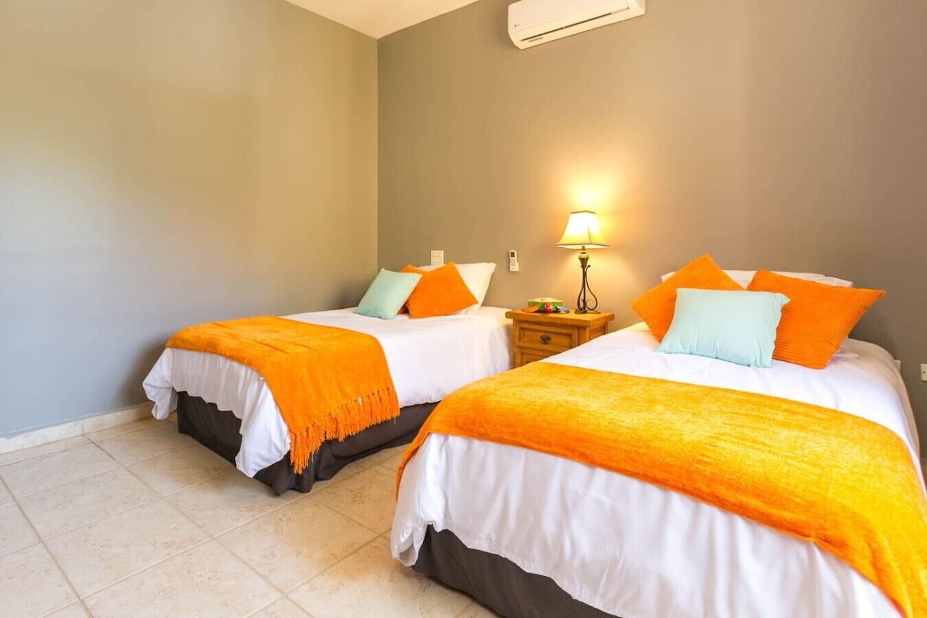 A hotel room with two beds, each with white bedding and orange throw blankets, a bedside table with a lamp, and a wall-mounted air conditioner.
