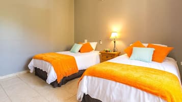 A hotel room with two beds, each with white bedding and orange throw blankets, a bedside table with a lamp, and a wall-mounted air conditioner.