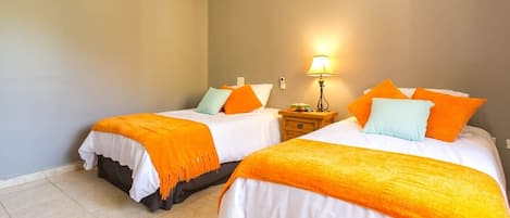 Une chambre d’hôtel avec deux lits, chacun doté de draps blancs et de plaids orange, une table de chevet avec une lampe et un climatiseur fixé au mur.