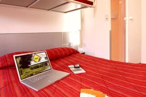 A bed with a laptop displaying a landscape, a bedside table with a cup, and a door in the background. - Premiere Classe Poitiers Futuroscope - Chasseneuil (Chasseneuil-du-Poitou)