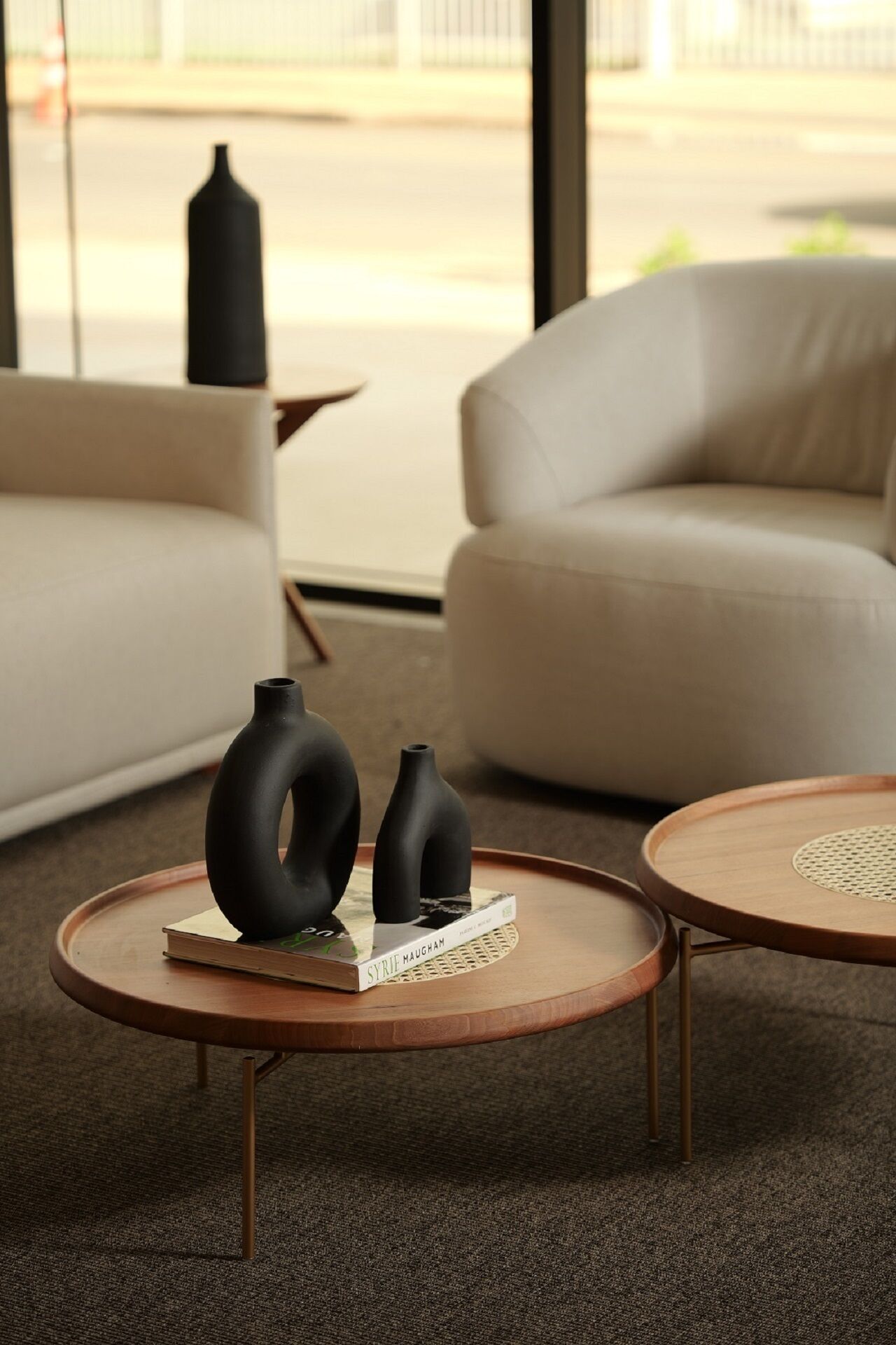 Un salon moderne avec deux tables rondes en bois, un canapé beige et deux vases noirs posés sur l’une des tables.