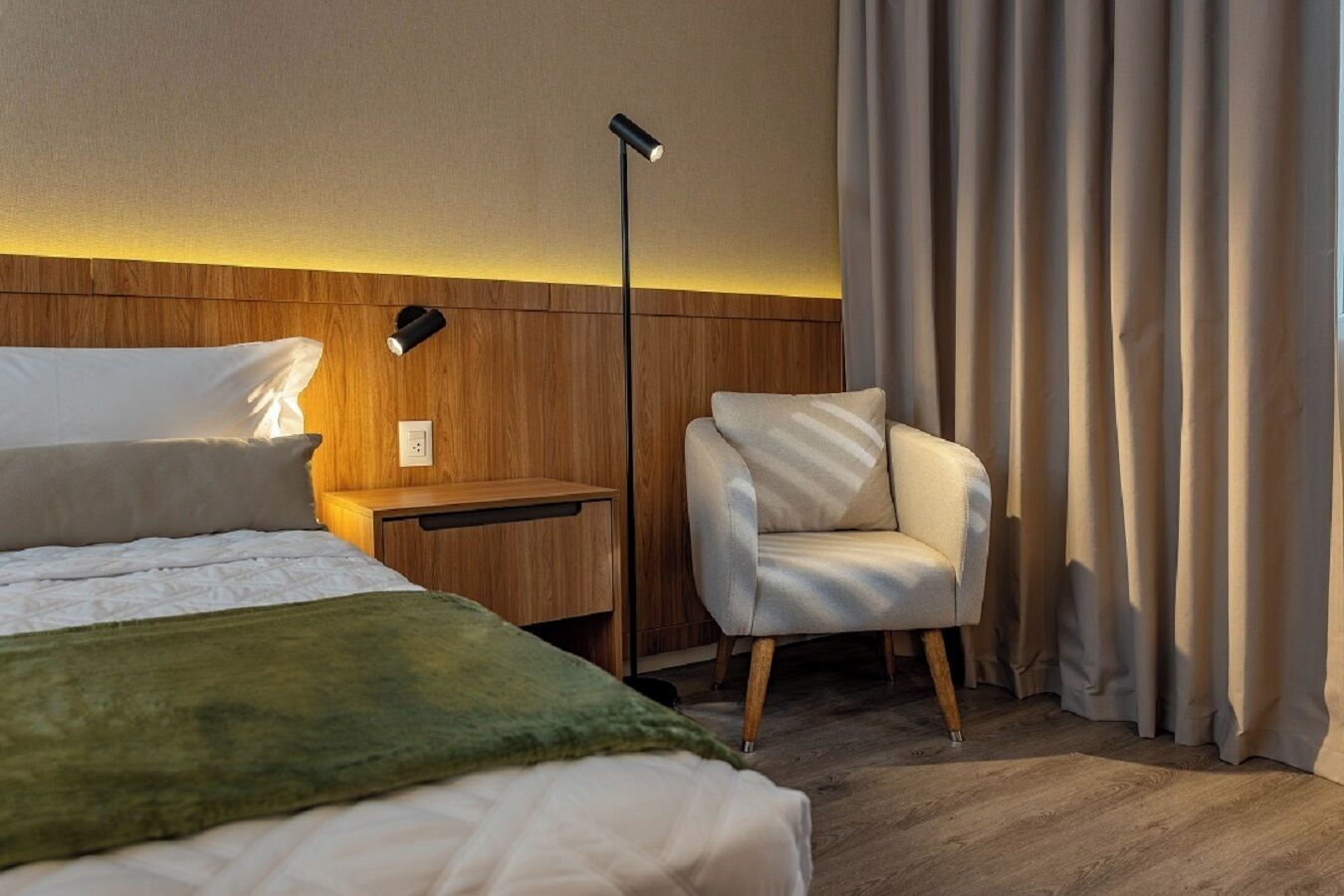 A hotel room with a bed, a nightstand, a chair, a floor lamp, and a curtain.