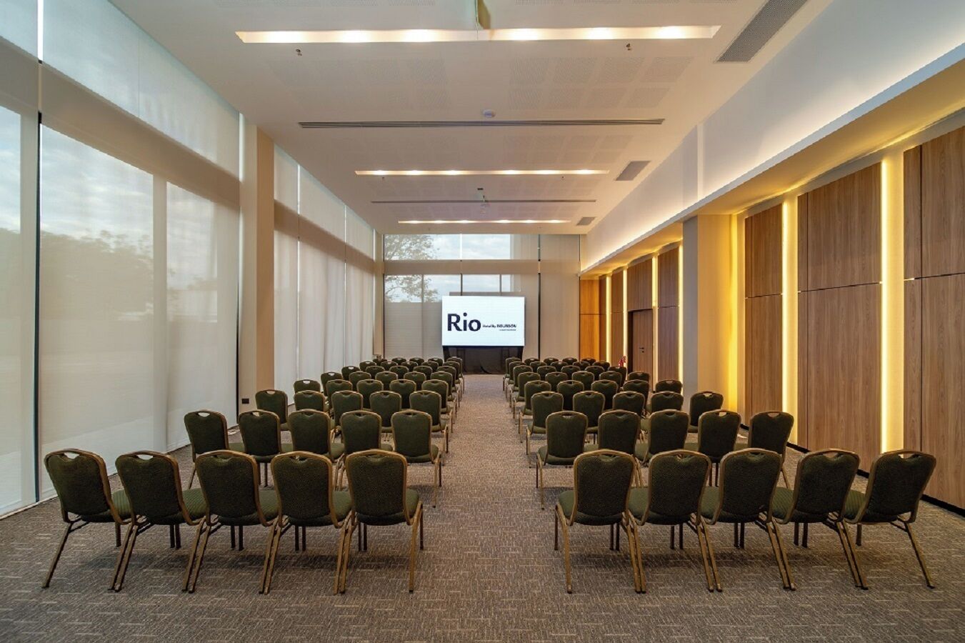 Una sala de conferències amb files de cadires orientades cap a una pantalla de projecció on es veu 'Rio'.