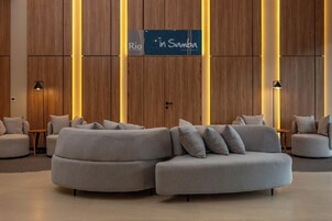Un vestíbulo moderno con un amplio sofá modular, paneles de madera y un letrero para el restaurante 'Rio in Samba'.