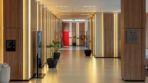 Un couloir d’hôtel moderne avec des panneaux de bois, des plantes en pot et une porte rouge au bout.