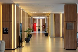 Un pasillo moderno de hotel con paneles de madera, plantas en macetas y una puerta roja al final.
