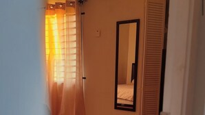 A narrow hallway with a tiled floor, a bed with a white mattress and a wooden headboard, a mirror on the wall, and a door with a sliding shutter.