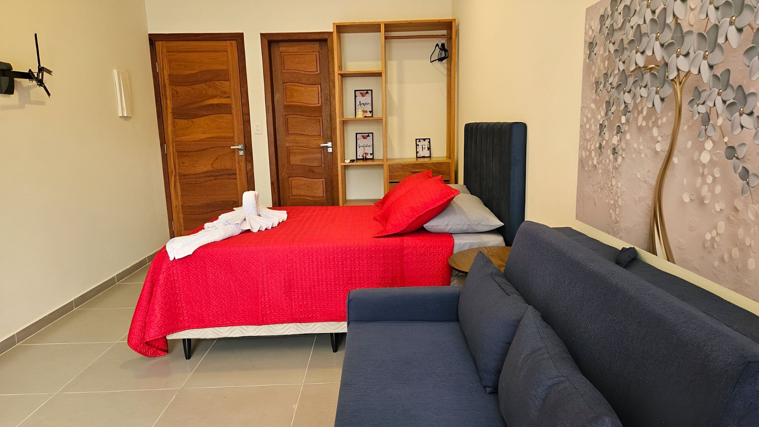 A hotel room with a bed, a sofa, a wooden wardrobe, a shelf with framed pictures, and a decorative wall panel.