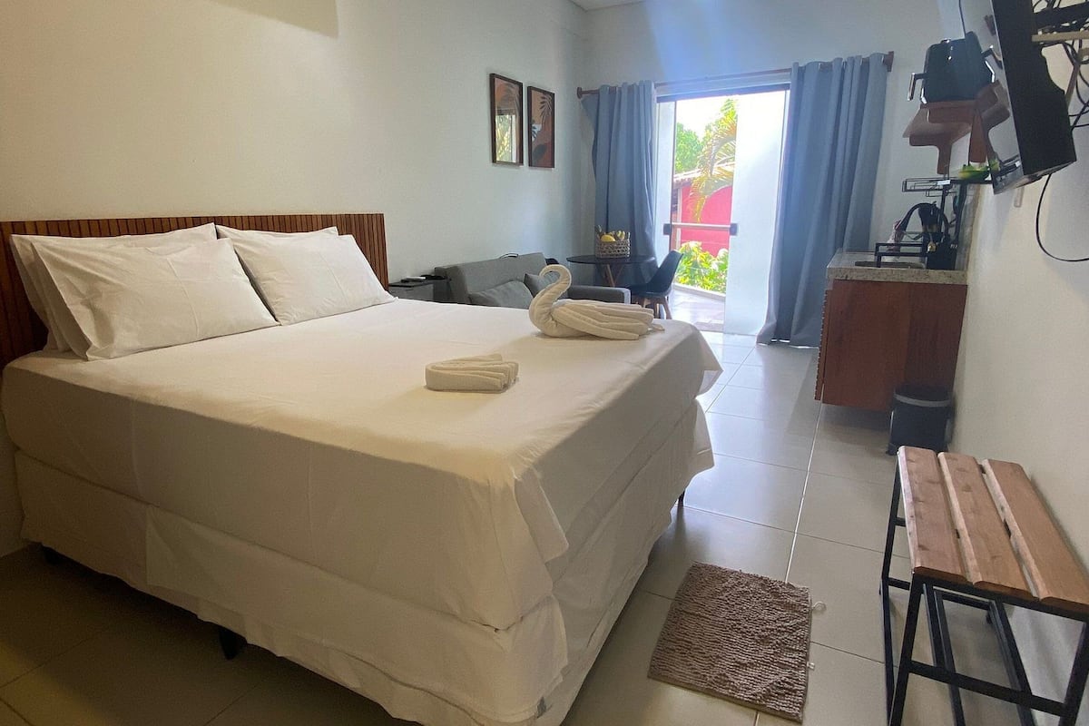 Quarto de hotel com cama, escrivaninha, cadeira, ar condicionado, ventoinha no teto e televisão.