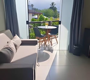 A modern living room with a sofa, a small round table, and chairs. There is a balcony with a view of greenery and buildings. - Residencial Mirante (Itacare)