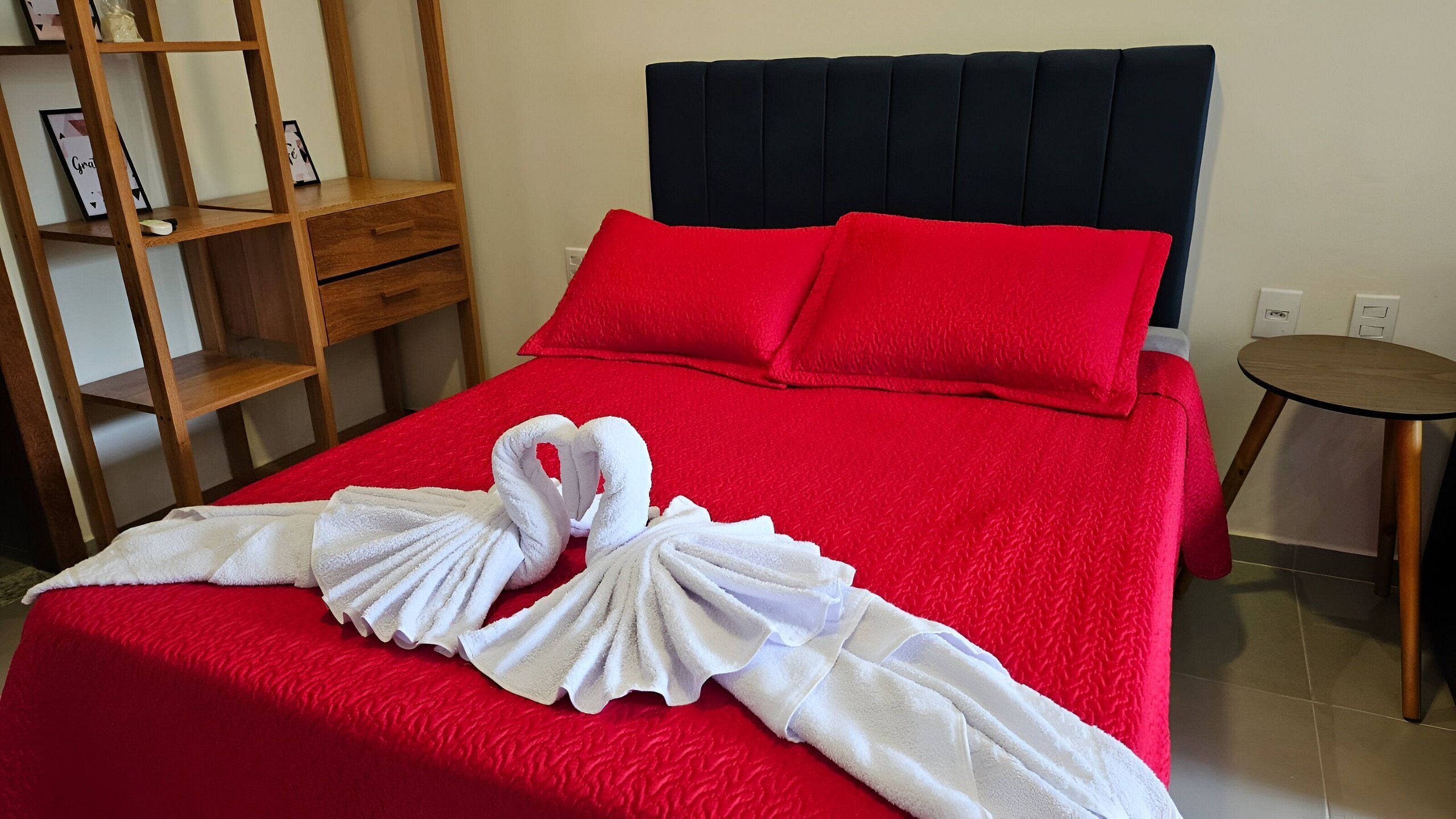 Ein Hotelzimmer mit rotem Bettbezug, zwei roten Kissen, einem Nachttisch aus Holz mit einem gerahmten Bild und einem kleinen Rundtisch.