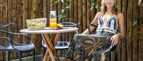 A person seated at a round table with a basket of fruit and a bottle of juice, outdoors with a bamboo fence and greenery in the background.