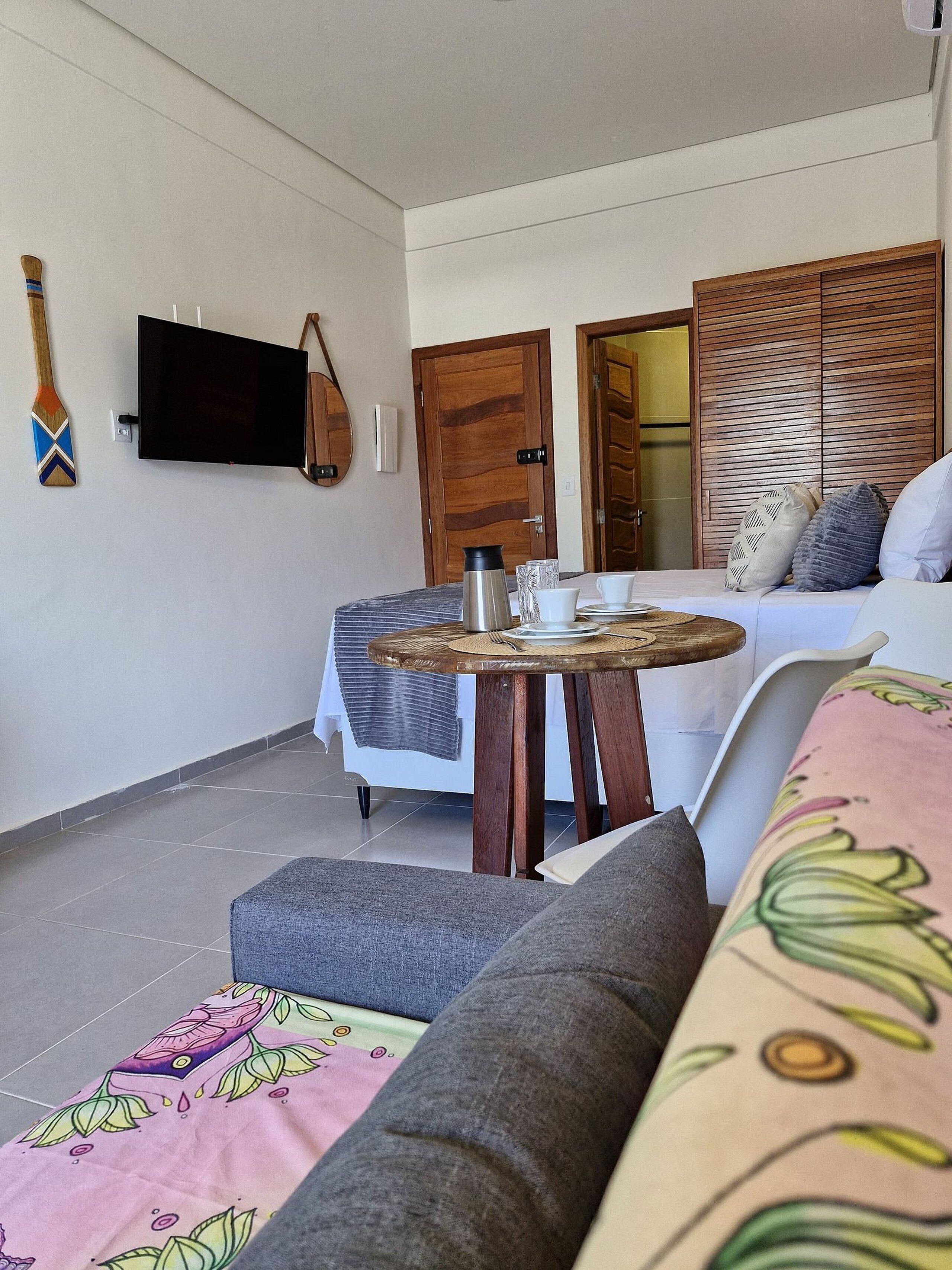 Une chambre avec un lit, une petite table ronde avec une bouilloire, un téléviseur fixé au mur et une porte en bois.
