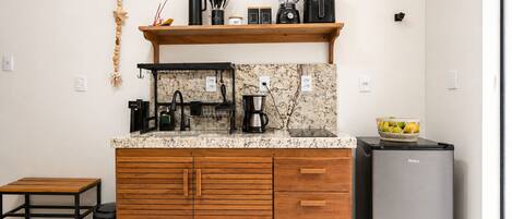 A compact kitchen with a marble countertop, wooden cabinets, a coffee maker, a blender, and a small refrigerator.