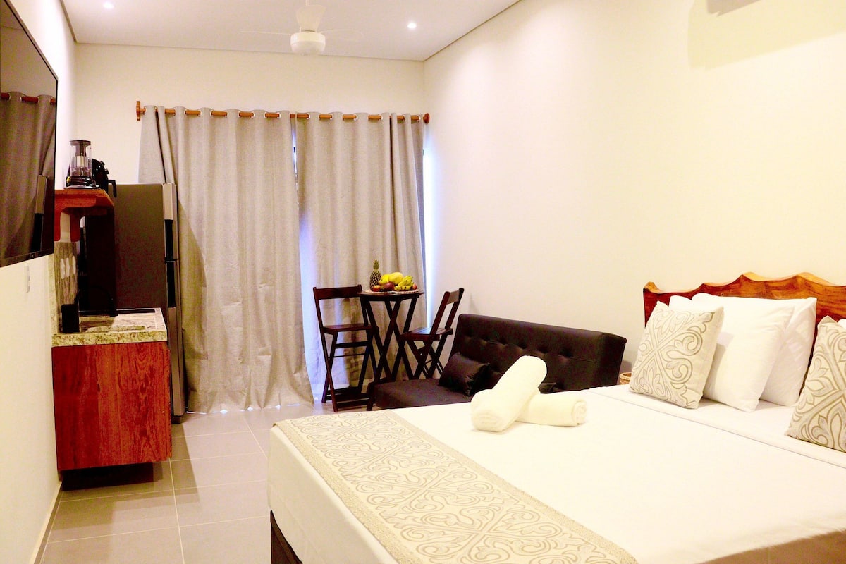 Quarto de hotel com cama, escrivaninha, cadeira, mesa com frutas e cortina.