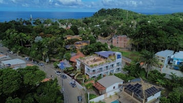 An aerial view of a residential area with houses, some with solar panels, surrounded by dense greenery and a view of the ocean.