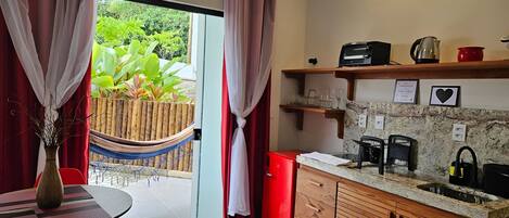 A modern kitchen with a red refrigerator, a coffee maker, and a kettle on the countertop. There is a wooden cabinet and a small shelf above the sink. A hammock is visible outside the window, and a table with chairs is in the foreground.