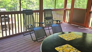 A covered outdoor area with a wooden deck, two chairs, a table, and a view of a forest. - Charming 3-bedroom cottage in Oxford with WiFi and AC close to Wisconson Dells (Oxford)