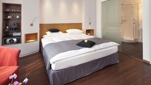 A modern hotel room with a large bed, a built-in shelf with kitchenware, a separate seating area, and a glass-enclosed shower. - Hotel Rutli (Zurich)