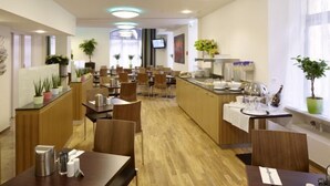 A modern hotel dining area with wooden flooring, multiple tables and chairs, a central counter with a glass display, and potted plants. - Hotel Rutli (Zurich)