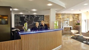 A reception desk with two staff members, a stone wall, and modern chairs in the background. - Hotel Rutli (Zurich)
