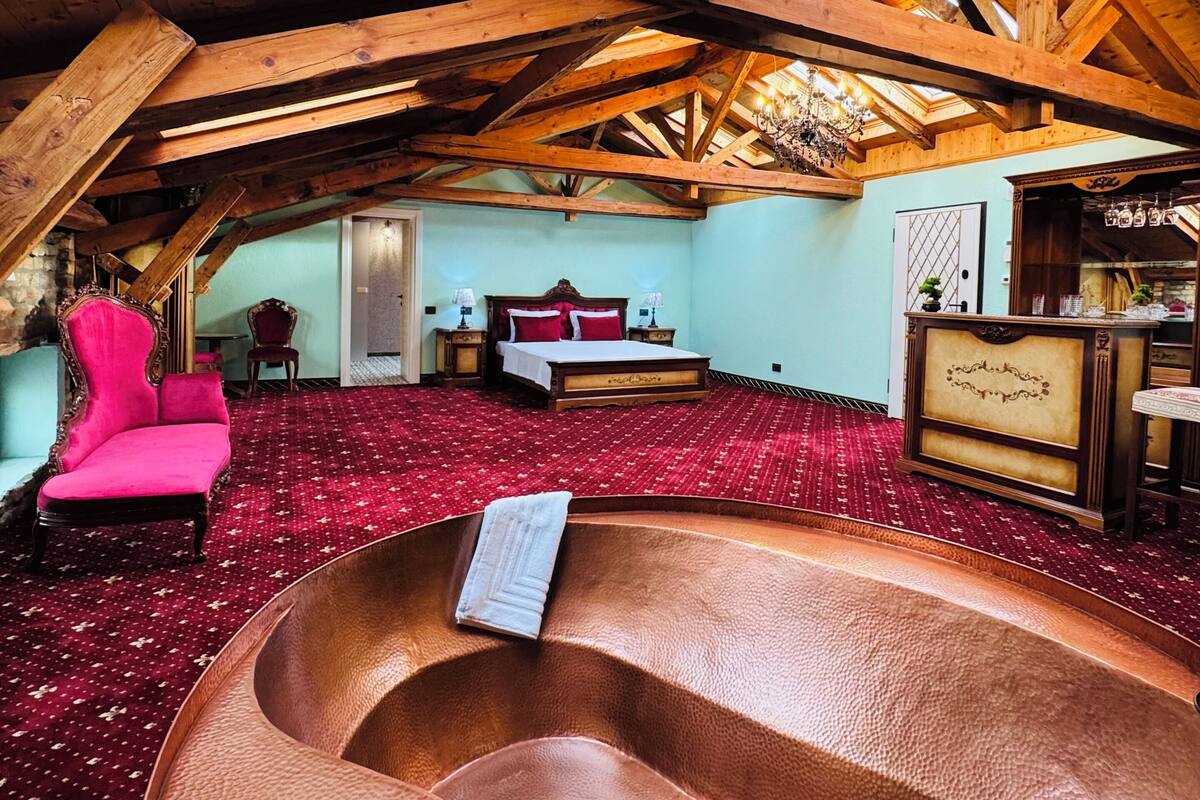 A spacious room with a high ceiling and wooden beams, a large bed with red bedding, a red carpet, and a unique copper bathtub.