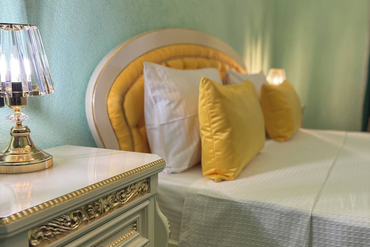 A neatly made bed with a white headboard and yellow pillows, a bedside table with a lamp, and a dresser with drawers.