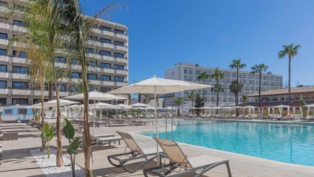 Un espace piscine doté de chaises longues et de parasols, entouré de palmiers et des bâtiments de l’hôtel.