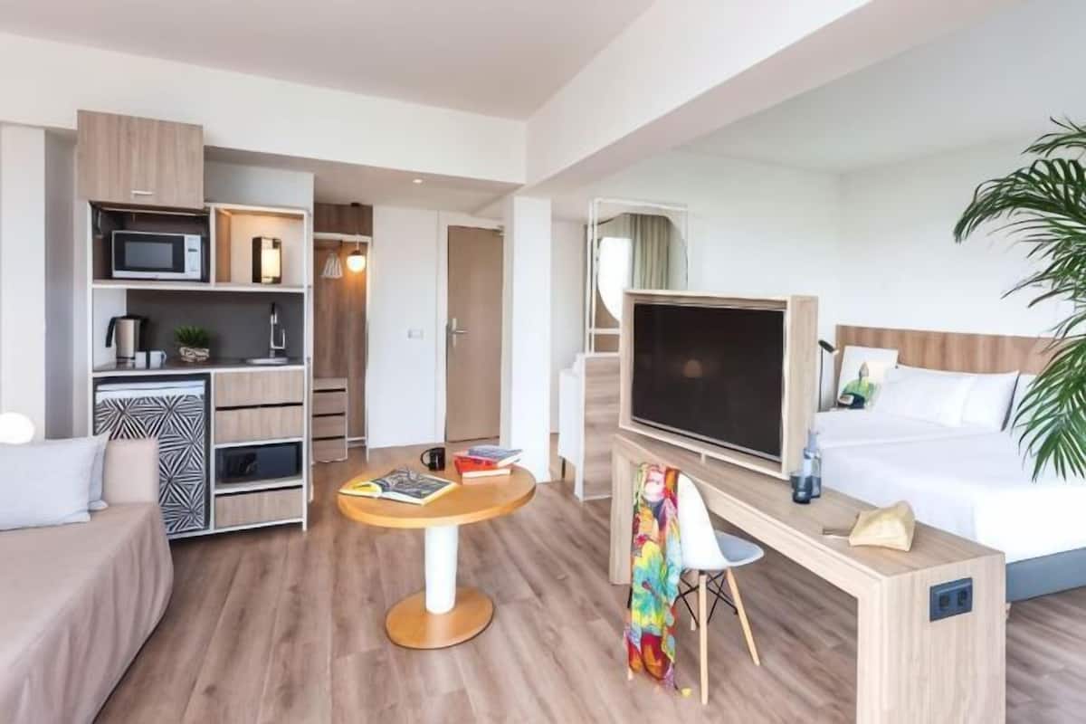 Quarto de hotel moderno com cama, televisão, escrivaninha e cozinha compacta.