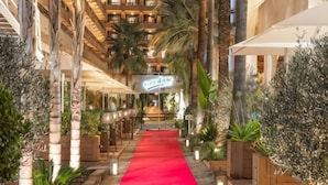Un couloir d’hôtel avec un tapis rouge, des palmiers et une enseigne indiquant « Hôtel de Rome ».