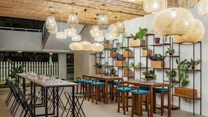 Un espace repas avec de longues tables et des chaises, des lampes en osier suspendues et des étagères ornées de plantes en pot.