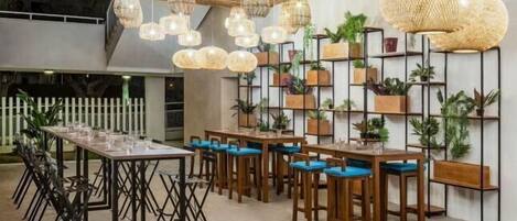 A dining area with long tables and chairs, hanging wicker lamps, and shelves with potted plants.