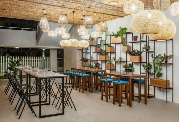 Un comedor con mesas largas y sillas, lámparas de mimbre colgantes y estantes con plantas en macetas.