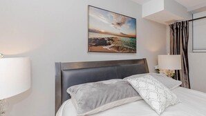 Ein Hotelzimmer mit einem Bett, einem Nachttisch, einer Lampe, einem Bild mit Strandmotiv und einem Fenster mit Vorhängen.