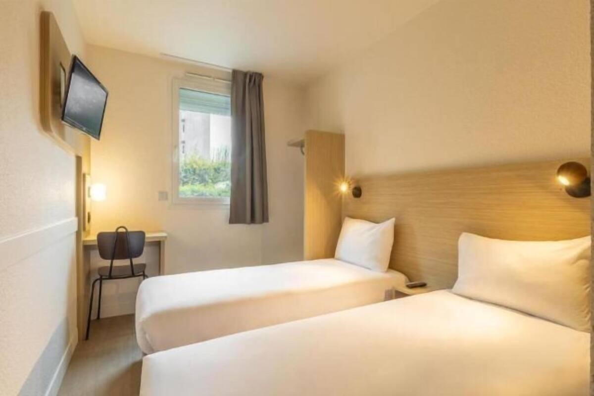 Ein Hotelzimmer mit zwei Betten, einem Schreibtisch, einem Stuhl, einem Fernseher und einem Fenster mit Vorhängen.