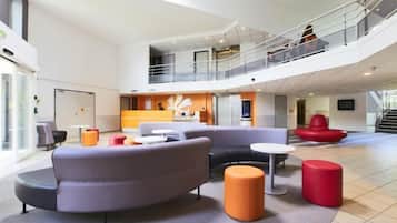 A spacious lobby with a high ceiling, a central seating area with sofas and ottomans, and a staircase leading to an upper level.