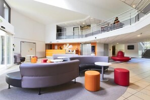 Un hall d’entrée spacieux avec un plafond haut, un espace de détente central agrémenté de canapés et de poufs, et un escalier menant à un niveau supérieur.
