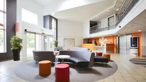 Un hall d’entrée spacieux, avec un canapé gris aux lignes courbes, une table ronde et un pouf rouge. Un escalier relie les deux niveaux, et une grande fenêtre laisse entrer la lumière naturelle.