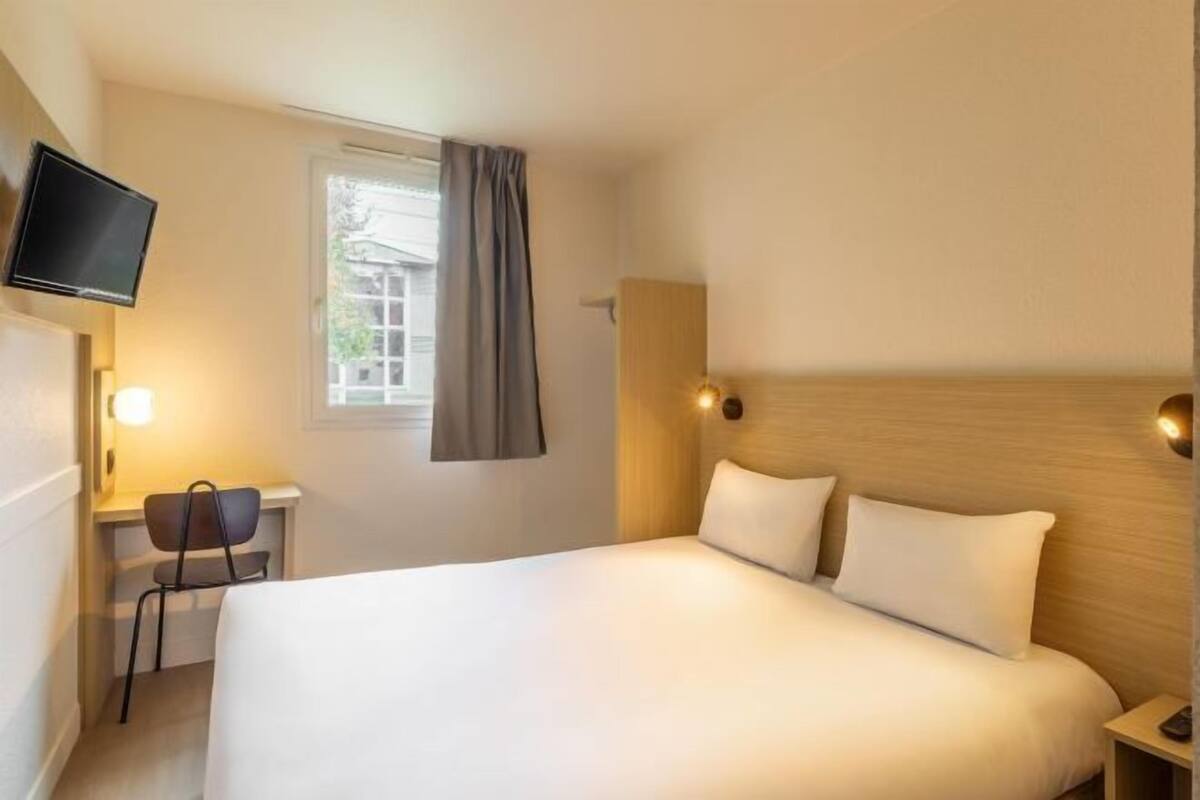 Ein Hotelzimmer mit einem Bett, einem Schreibtisch mit Stuhl, einem Fernseher und einem Fenster mit Vorhängen.