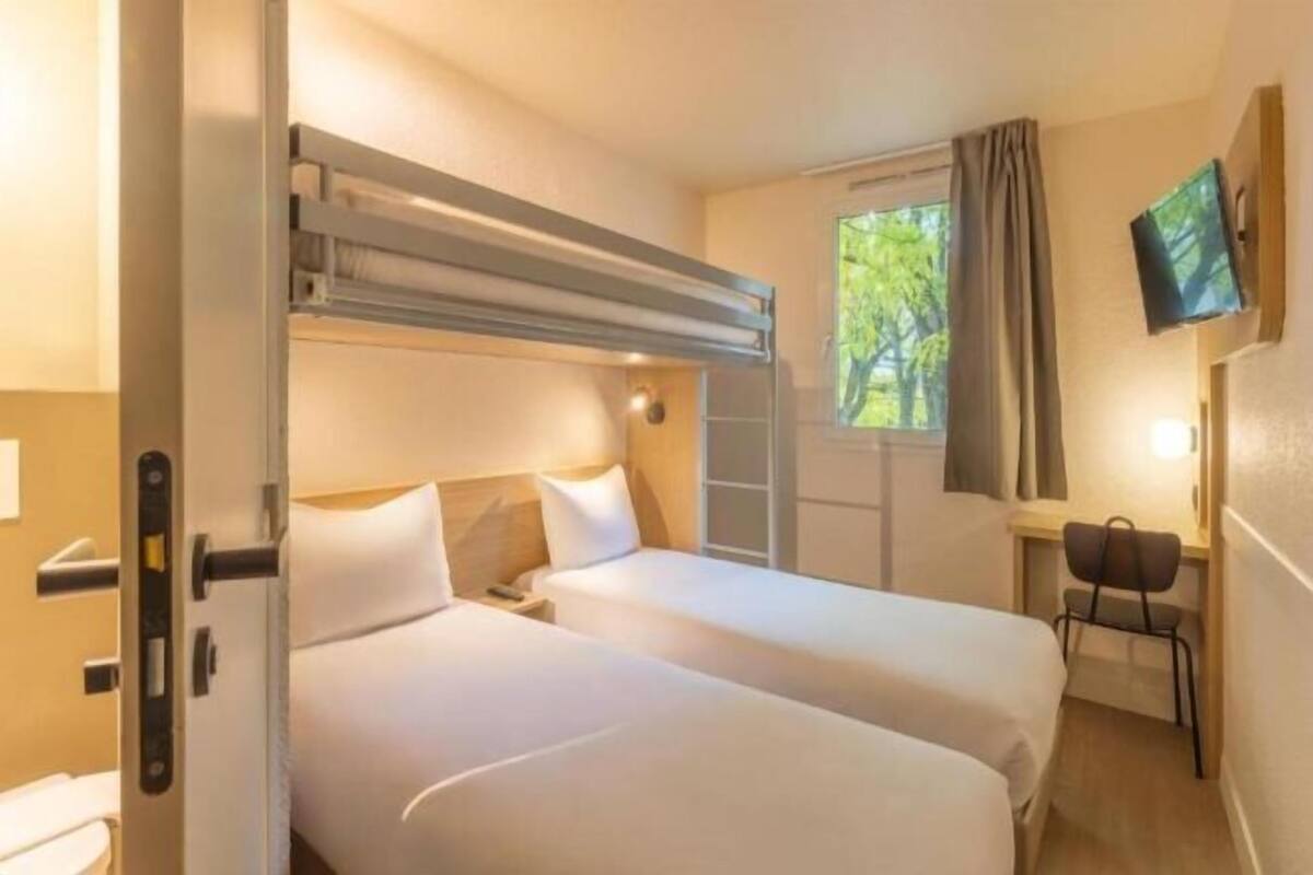 Ein Zimmer mit zwei Betten, einem Schreibtisch, einem Stuhl, einem Fernseher und einem fenster mit Vorhängen.