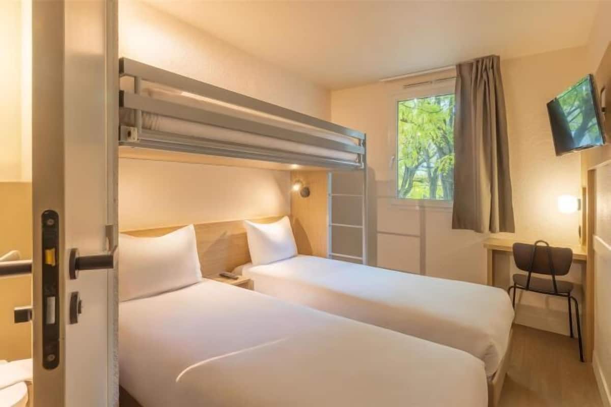 Ein Hotelzimmer mit zwei Betten, einem Schreibtisch mit Stuhl, einem Fernseher und einem Fenster mit Vorhängen.