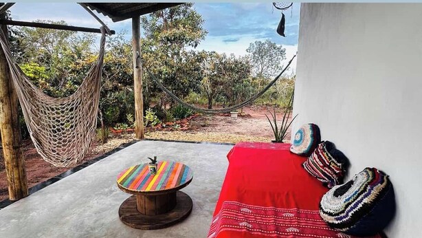 Un hamac, un tabouret coloré, un canapé rouge et des coussins à motifs, le tout dans un décor extérieur avec vue sur les arbres et un ciel dégagé.