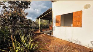 Un bâtiment blanc avec une porte et une fenêtre en bois, entouré de végétation et doté d’un porche couvert.