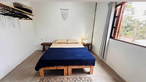 Une petite chambre à coucher accueillante, avec un lit simple, une table de chevet, une étagère fixée au mur et une fenêtre avec des rideaux.