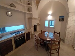 A kitchen with a round wooden table, a built-in oven, and a window above the fireplace. - "Al Vicolo della Colonna" Vacation Home, Historic Center of Presicce (Presicce)