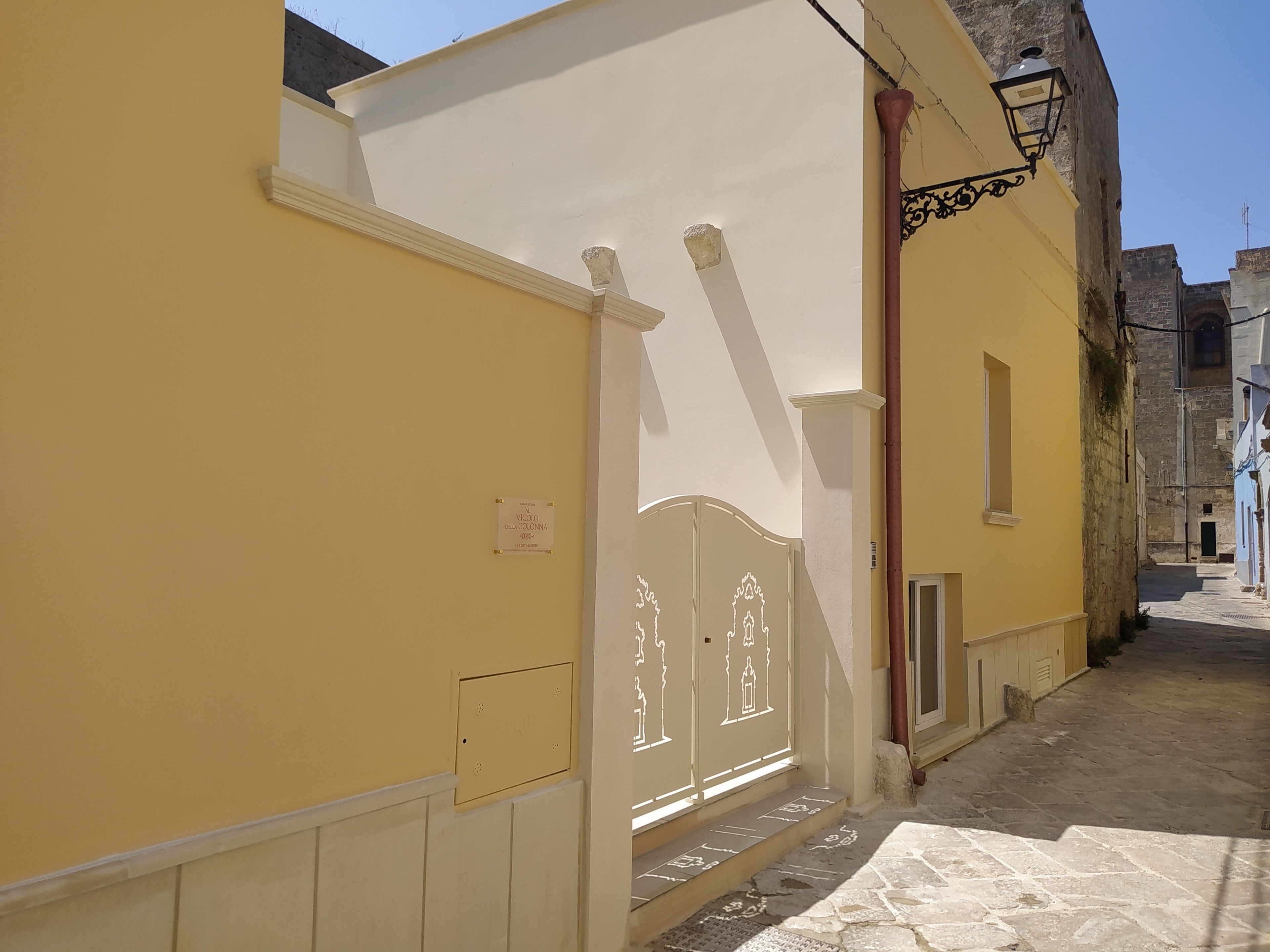 Une rue étroite bordée d’un immeuble jaune, d’un portail blanc et d’un chemin pavé de pierres.