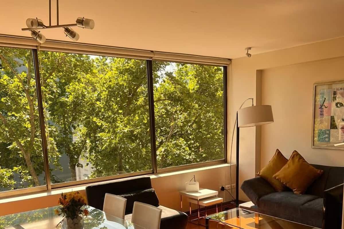 A modern living room with a glass dining table, a sofa, and a large window offering a view of trees.
