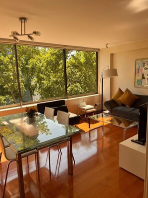 Uma sala de estar moderna com uma mesa de jantar de vidro, um sofá e uma grande janela com vista para árvores.