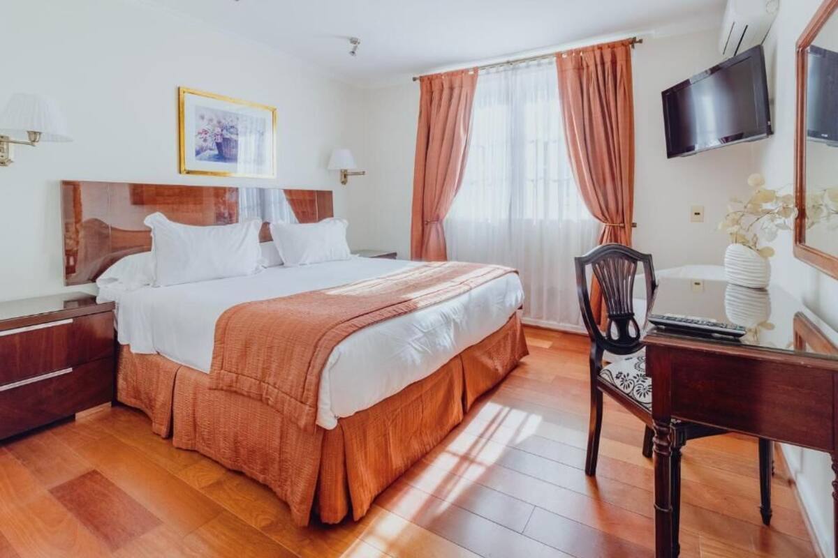 A hotel room with a wooden floor, a bed with white bedding, a wooden headboard, a desk with a chair, a television, and a window with curtains.